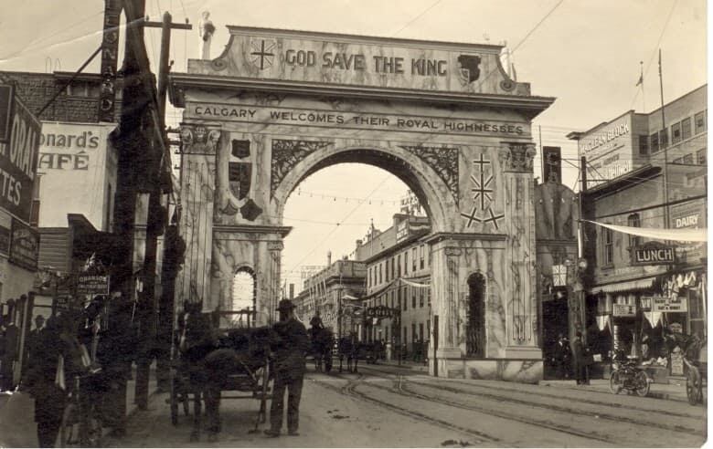 In a black and white photo, a high archway straddles a downtown street. Across the top of the arch it reads God save the King.