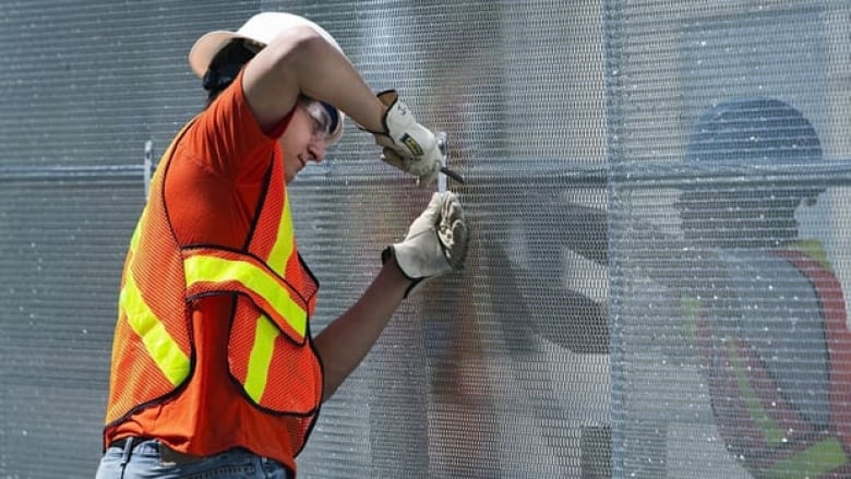 A man in an orange safety vest with white gloves and white hardhat works with a tool on some fencing