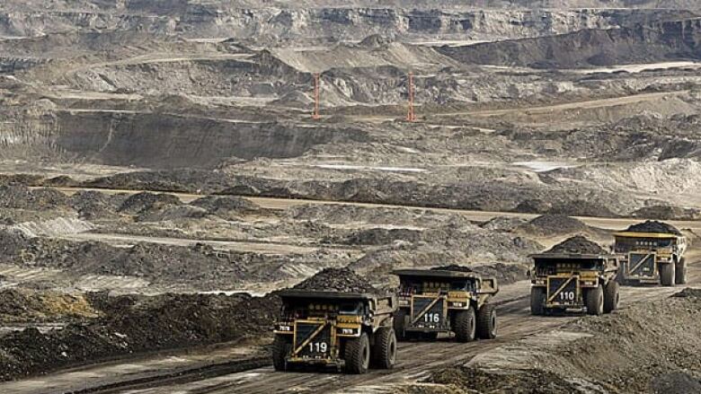 A row of large mining trucks travel down a dirt road in the oilsands.