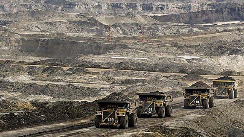 A row of large mining trucks travel down a dirt road in the oilsands.