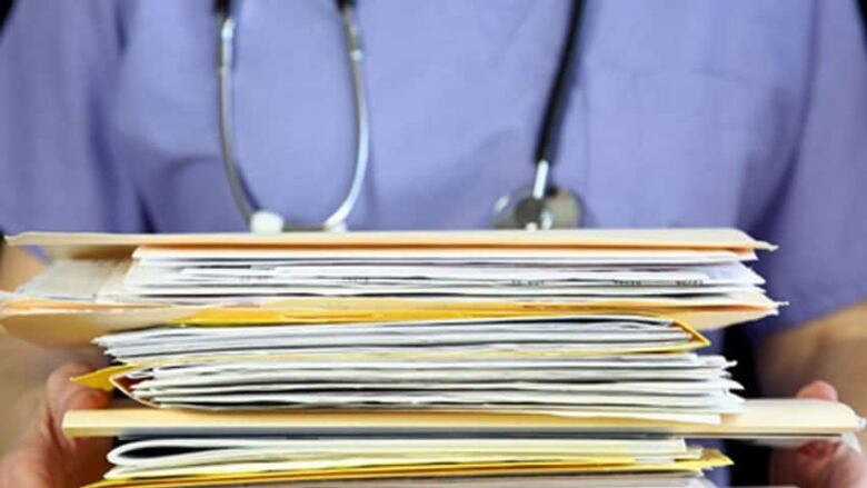 A stack of medical files are held by someone wearing blue scrubs and a stethoscope.