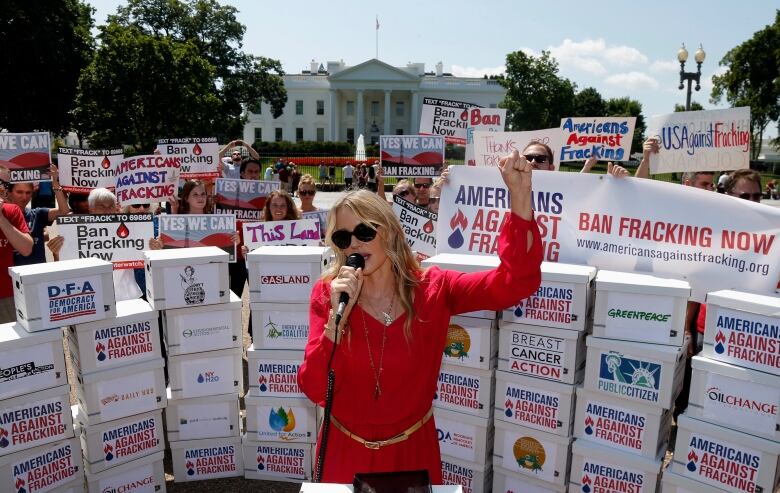 A woman in a red dress stands, left fist raised and right hand holding a microphone, surrounded by filing boxes and protesters with anti-fracking signs.