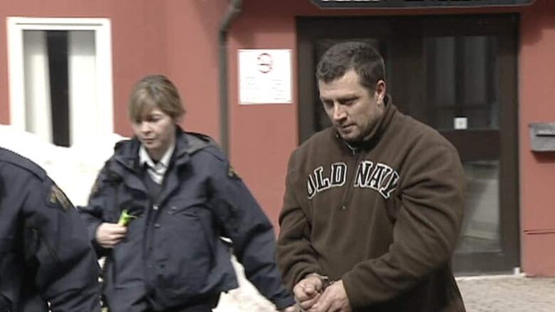 A man wearing a hoodie and restrained in handcuffs is led away from a courthouse by a female security officer in uniform.