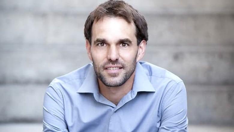 A man wearing a light blue dress shirt looks at the camera. He has brown hair and stubble. 