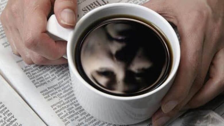 Reflection of a woman's face in a cup of black coffee which is sitting on top of a newspaper.