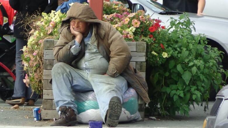 A man in an open winter coat sits on the sidewalk in front of a flower box. 