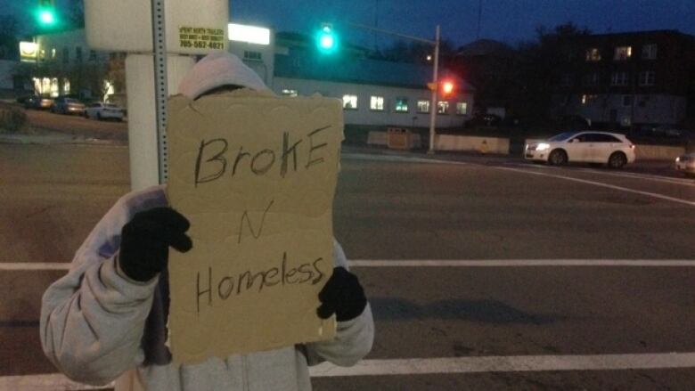 A man wearing a hoodie standing at a busy intersection holds a sign up covering his face. It reads 