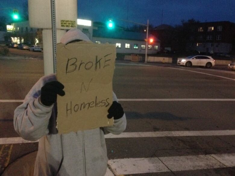 A man wearing a hoodie standing at a busy intersection holds a sign up covering his face. It reads 