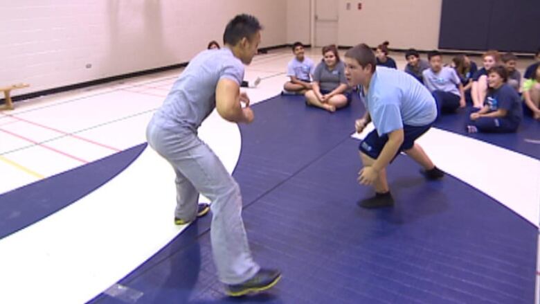 A staff member coaches students in wrestling on a blue mat in a gym.