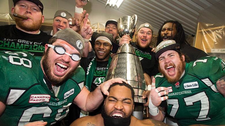 A group of men cheer and make faces while standing and sitting around a giant silver cup.