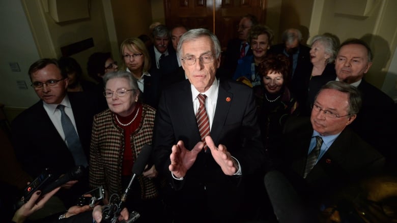 A man wearing a suit, surrounded by other people, speaks to people holding microphones.