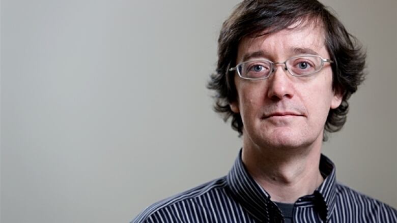 A man wearing glasses and a collared shirt poses for a photo in front of a grey background.