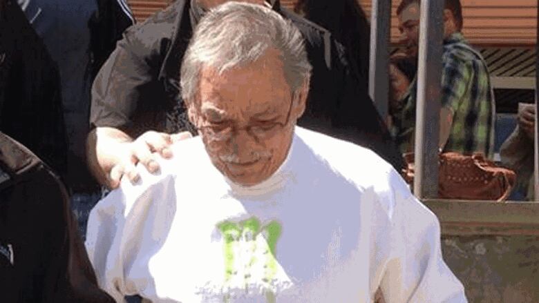 A portrait of a man with grey hair and moustache, wearing glasses and a white sweatshirt, with his head down and the hand of a supporter behind him on his right shoulder.