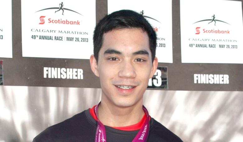 A young man with dark hair smiles while holding up a medal around his neck that says 