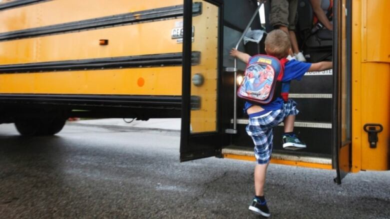 A little boy wearing a Spider-Man backpack steps onto a yellow school bus.