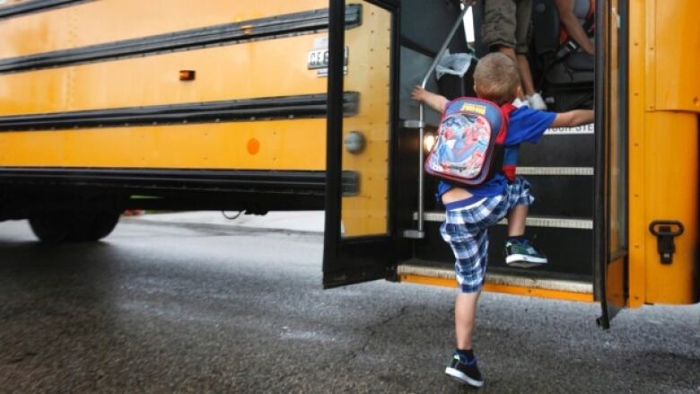 A little boy wearing a Spider-Man backpack steps onto a yellow school bus.