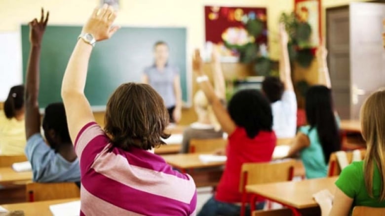 A classroom of students is seen from behind, with the students raising their hand to answer a teacher in the front of the classroom.