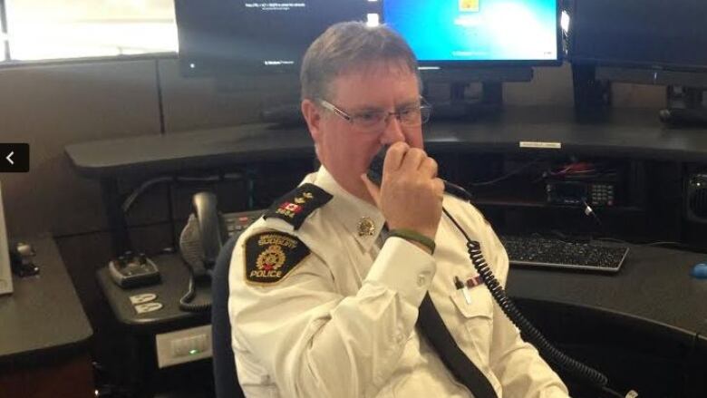 Todd Zimmerman speaks into a police radio, sitting at a desk with several computer screens, wearing a white police uniform