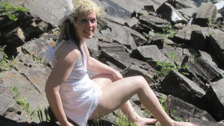 A woman wearing a white dress sits on a rock pile near the ocean. 