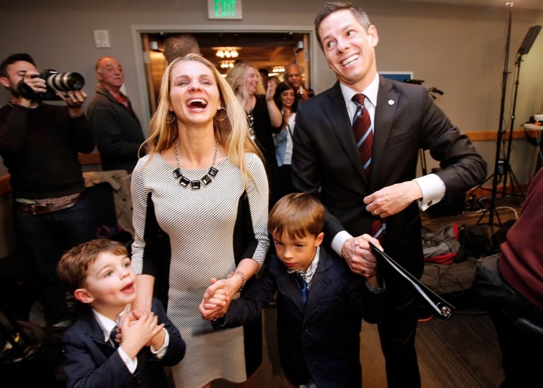 With a news photographer in the background, a smiling man in a suit stands to the left of a laughing woman in a dress. They hold the hands of a young boy standing between them, while the woman holds the hand of another young boy on her right.