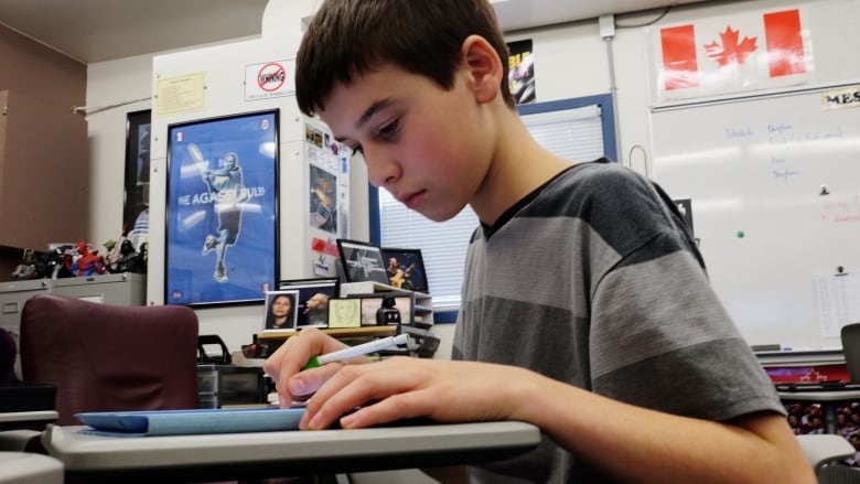 A young boy looks down at an electronic device on his desk in a school room setting.