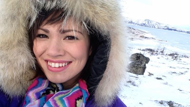 A smiling woman wearing a purple parka with a fur-lined hood stands outside, with a snowy expanse, with a body of water and hills behind her.