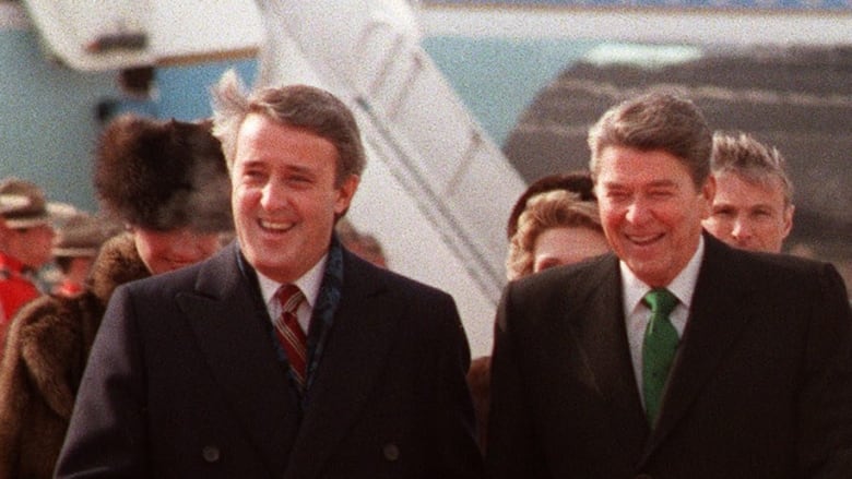 Two men walk down a red carpet from an airplane as a line of men in red uniforms stand at attention.