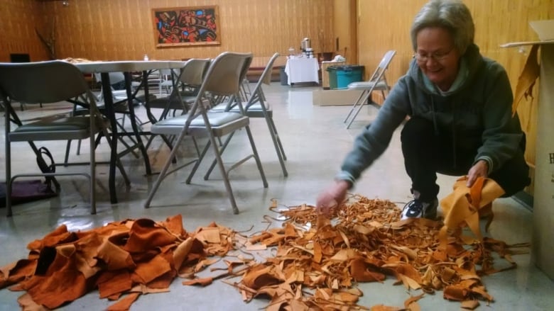 A woman sorts through a pile of tan and brown leather strips scattered on the floor.