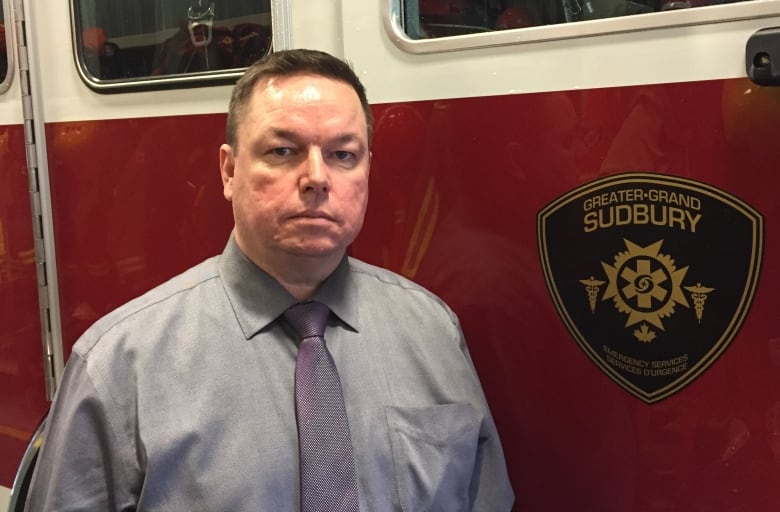 A straight-faced man in a shirt and tie stands next to a Sudbury fire truck