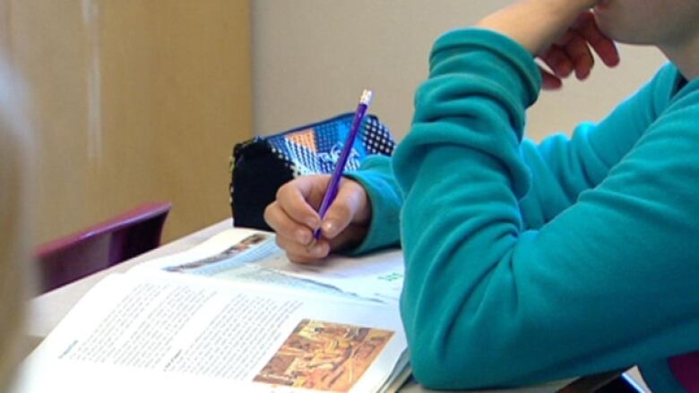 A student with a book open pauses in thought as they write on a piece of paper.