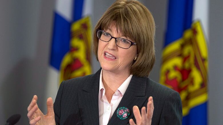 A woman with brown hair and glasses speaks at a podium, holding her hands up to explain a point. 