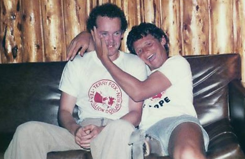 Two men sitting on a black couch. The man on right has his hand on the man on the left's cheek.