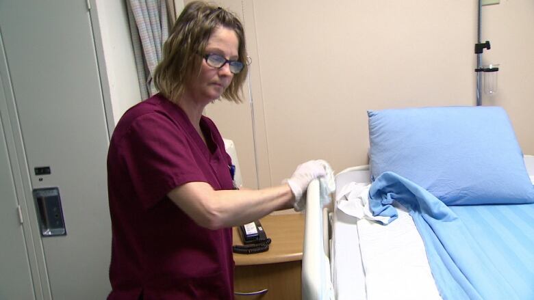 A health-care worker with shoulder-length brown hair and glasses, wearing burgandy scrubs and white medical gloves, wipes down a hospital bed railing. 