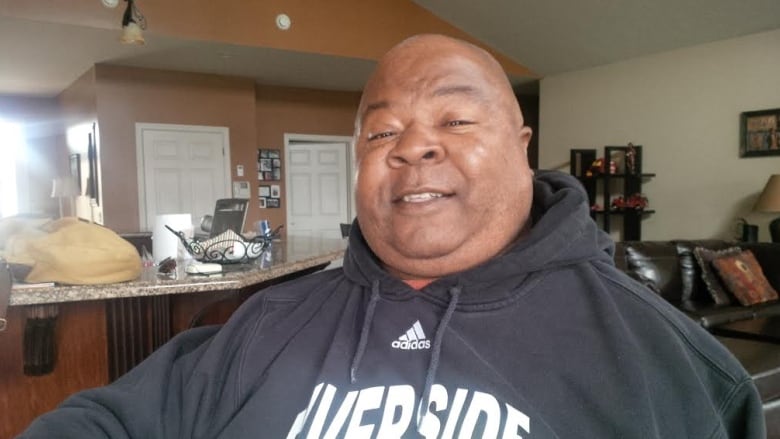 Head and shoulders shot of George in what appears to be an open concept home with a kitchen counter on one side of him in the background and a living room sofa on the other. 