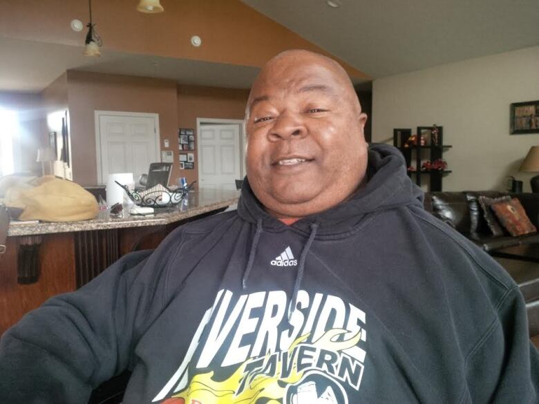 Head and shoulders shot of George in what appears to be an open concept home with a kitchen counter on one side of him in the background and a living room sofa on the other. 