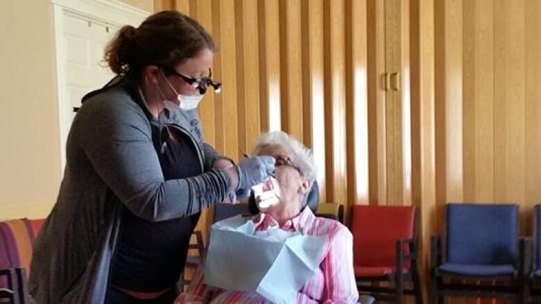 A woman in a black shirt and grey sweater performs a dental procedure on a woman in a pink shirt sitting on a medical chair. 