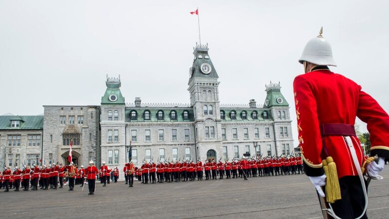 Officer cadets in red uniforms, black pants and white helmets are shown parading in front of a large, ornate brick building.