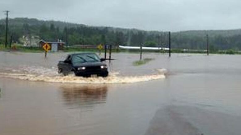 A pickup truck moves through a fully flooded road, creating a wake in its path.