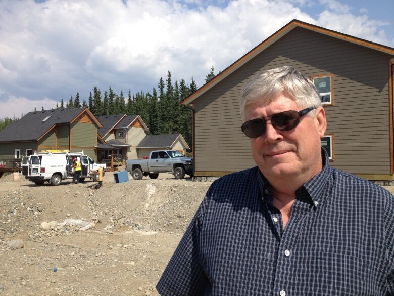 A man with white hair and glasses stands in front of some newly finished houses.