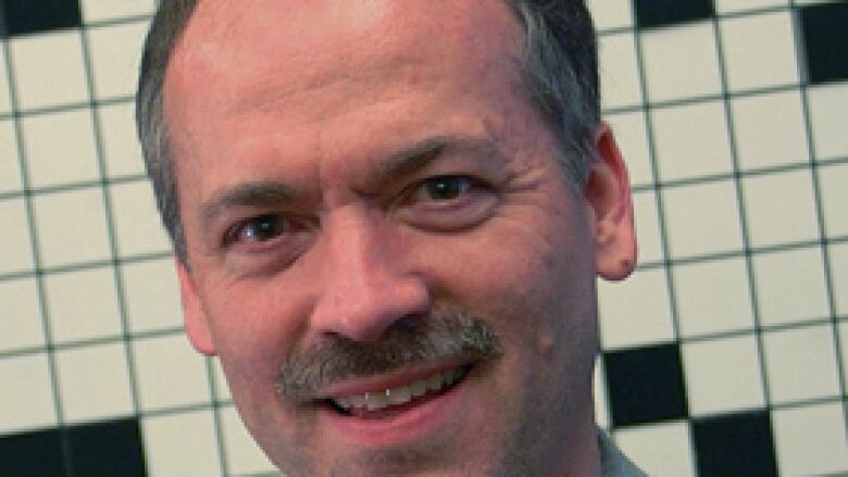 A headshot of a man in a light gray button up shirt. In the background is a blank crossword puzzle.