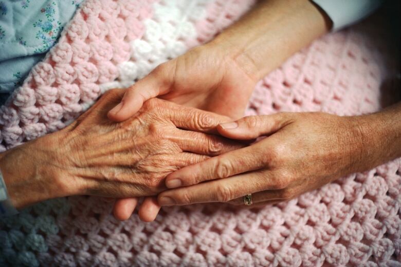 A person holds the hand of an elderly woman, who is covered with a pink and white crocheted blanket.