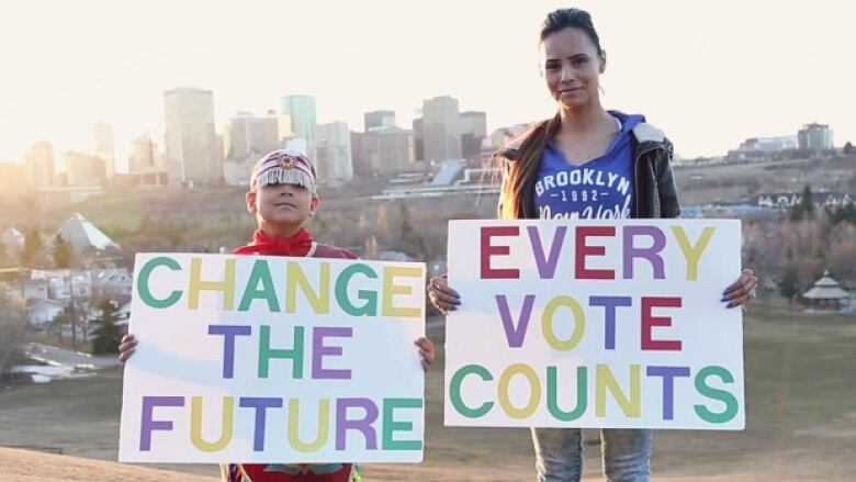 A woman and boy with the Edmonton skyline behind them hold signs reading 