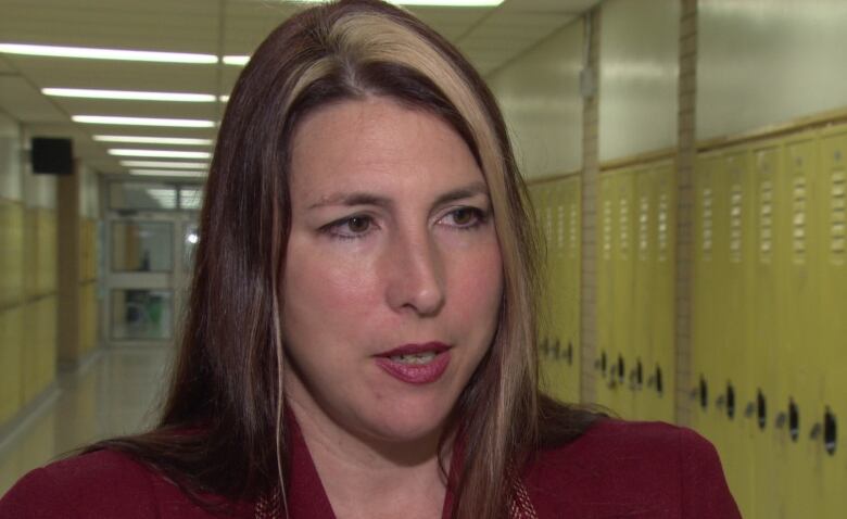 A woman with dark brown hair and chunky blonde highlights is speaking in a locker-lined school hallway.