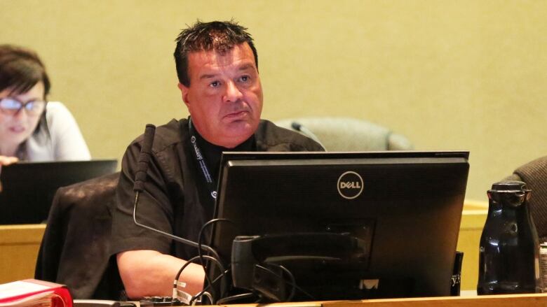 A man with black hair, wearing a black shirt sits at a table.