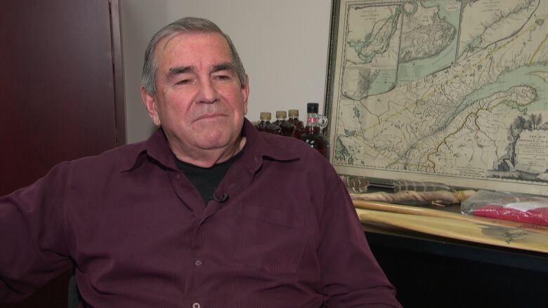 A man with grey hair and a maroon shirt on sits in front of a map.