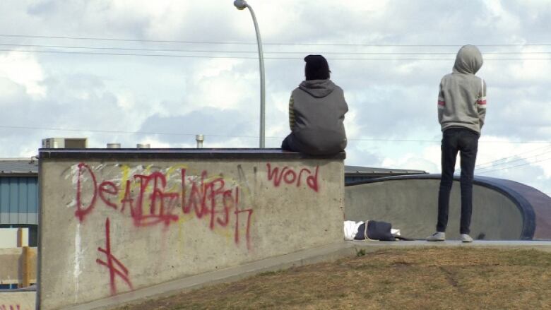 One child sits on a graffitied concrete block with another one standing nearby. Both have their backs to the camera.