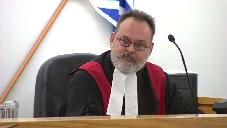 A man with a grey beard, glasses and wearing black and white robes sits at a bench with a microphone in front of him.