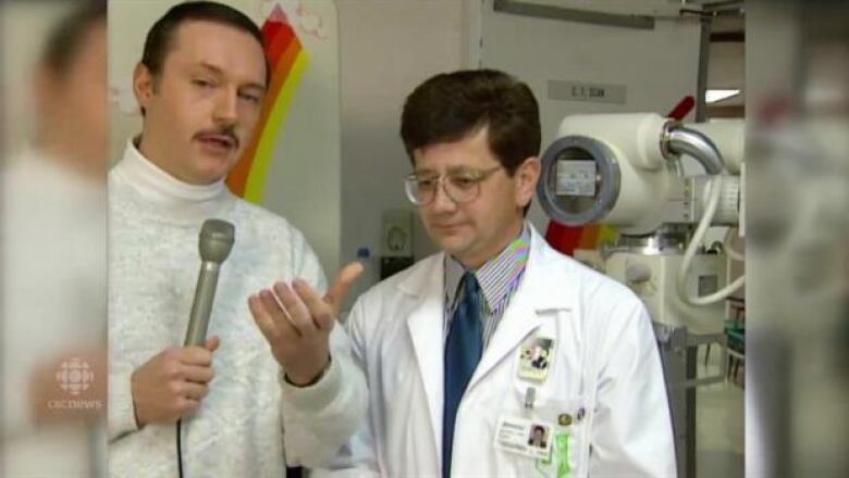 A man holding a microphone interviews a man wearing a white lab coat. 