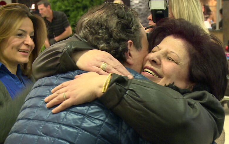 A man and woman embrace, the woman is smiling with her eyes closed while another woman looks on smiling.