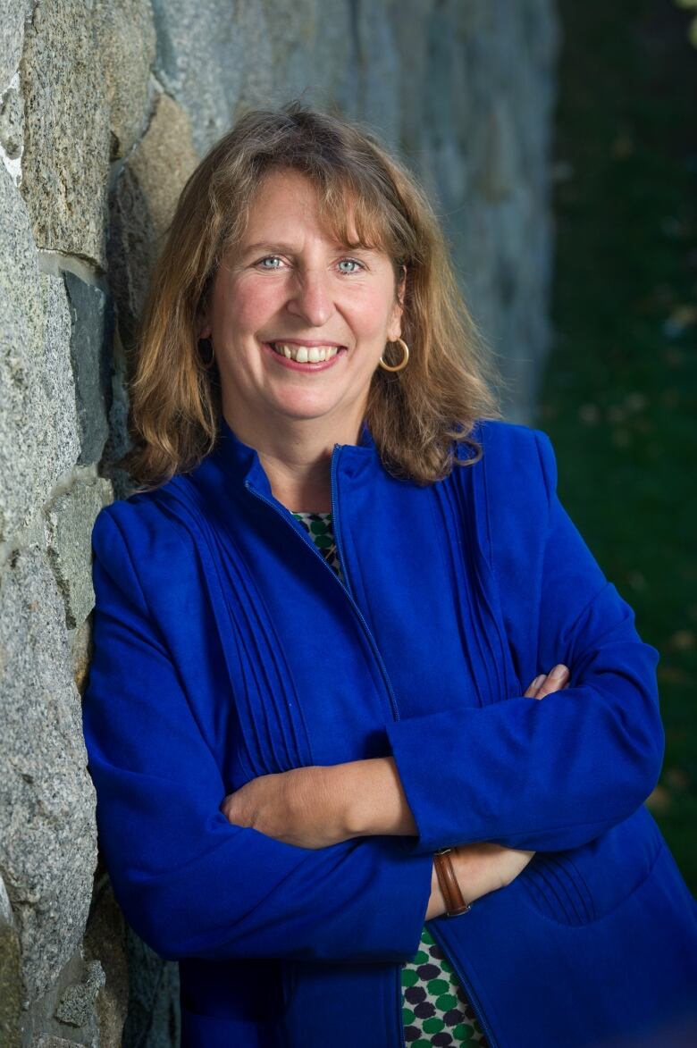 Person wearing blue, leaning against a wall, looking at the camera. 